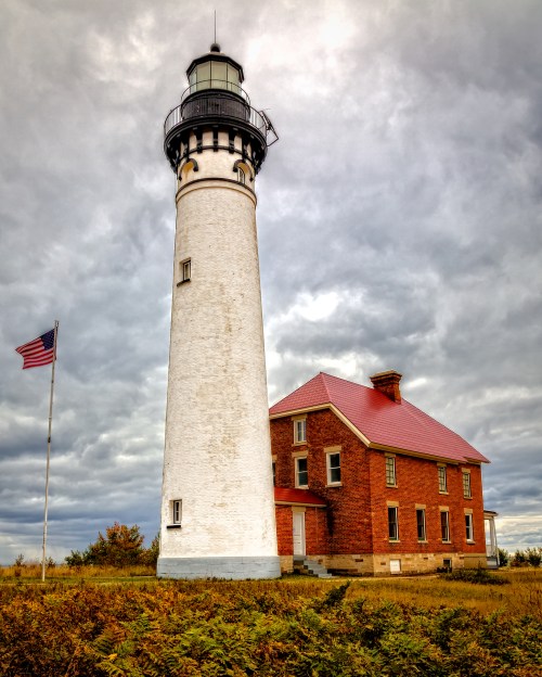 A tall white lighthouse beside a red-roofed brick house, with an American flag flying in front, under a cloudy sky.