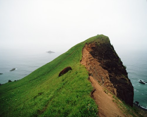 A grassy cliff edge overlooking a calm sea, with a small island visible in the distance under a cloudy sky.