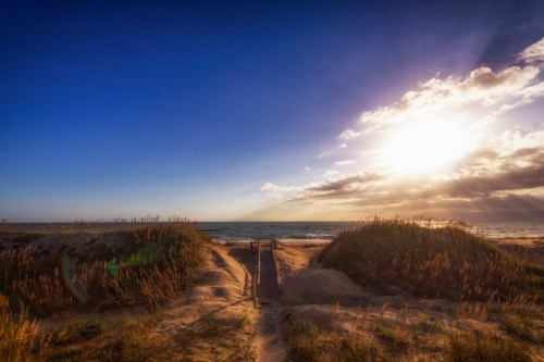 A sandy path leads to the ocean under a bright sky with clouds and a setting sun.