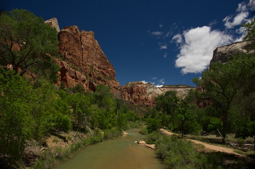 A serene river flows through a canyon, surrounded by towering red rock formations and lush green trees under a blue sky.