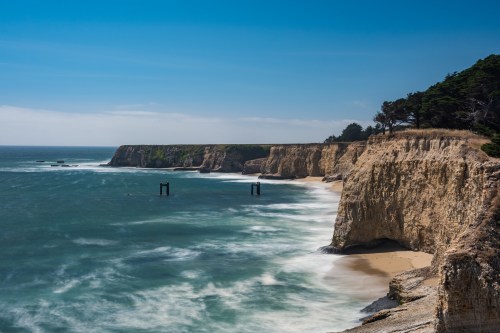A scenic coastal view featuring cliffs, a sandy beach, and gentle waves under a clear blue sky.