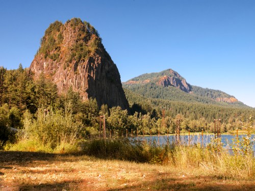Scenic view of two mountains beside a calm lake, surrounded by lush greenery under a clear blue sky.