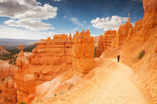 A person walks along a winding path through orange rock formations under a blue sky with scattered clouds.