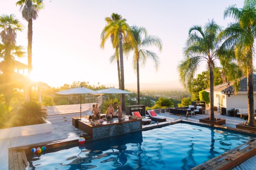A sunny poolside scene with friends enjoying drinks, surrounded by palm trees and a scenic view at sunset.