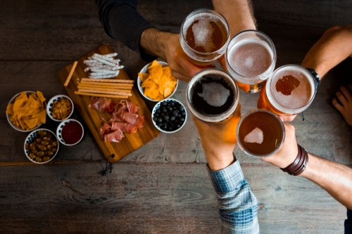 A group of hands toasting with glasses of beer over a table with snacks like chips, olives, and cured meats.