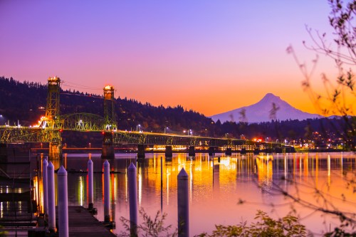 A vibrant sunset over a bridge with lights reflecting on the water, with a mountain in the background.