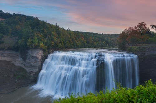 A stunning waterfall cascades over rocky cliffs, surrounded by lush greenery and colorful autumn trees under a pastel sky.