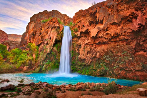A stunning waterfall cascades over rocky cliffs into a vibrant blue pool, surrounded by lush greenery and rugged terrain.