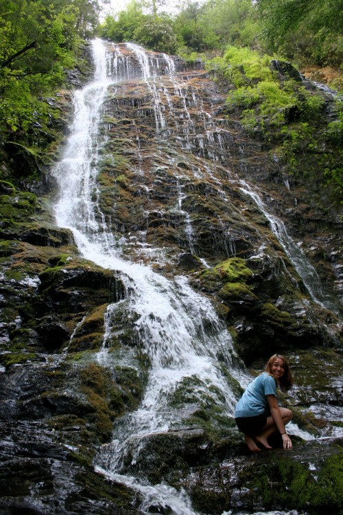 A person crouches by a cascading waterfall surrounded by lush greenery and rocky terrain.