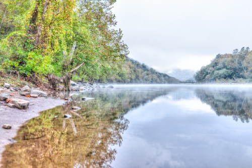 A serene river scene with mist, surrounded by lush greenery and rocky shores, reflecting the calm water.