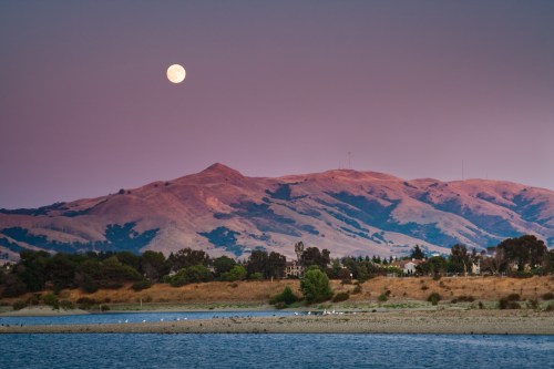 A serene landscape featuring a full moon over rolling hills, with a calm lake in the foreground and trees along the shore.