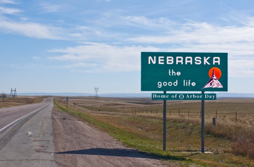 A green roadside sign reads "Nebraska the good life, Home of Arbor Day," with a scenic landscape and blue sky in the background.