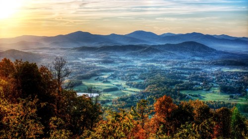 A panoramic view of rolling mountains at sunset, with vibrant autumn foliage and a misty valley below.