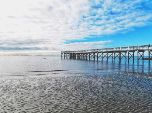 A wooden pier extends over calm water under a cloudy blue sky, with rippled sand in the foreground.