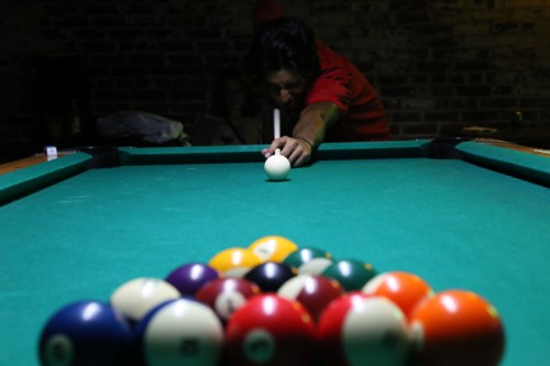 A person in a red shirt prepares to shoot a cue ball at a pool table with a triangle of colored balls.