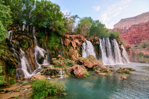 A scenic view of waterfalls cascading over rocks, surrounded by lush greenery and red rock formations.