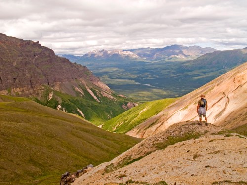A hiker stands on a rocky ledge, overlooking a vast green valley surrounded by mountains under a cloudy sky.