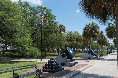 A park scene featuring cannons, cannonballs, and palm trees, with an American flag in the background under a clear blue sky.