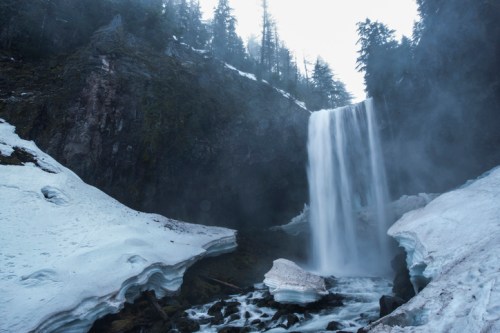 A majestic waterfall cascades over rocky cliffs, surrounded by snow and misty forest in a serene landscape.