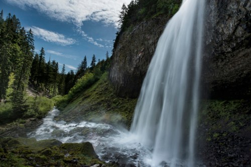 A majestic waterfall cascades down rocky cliffs, surrounded by lush green trees and a clear blue sky.