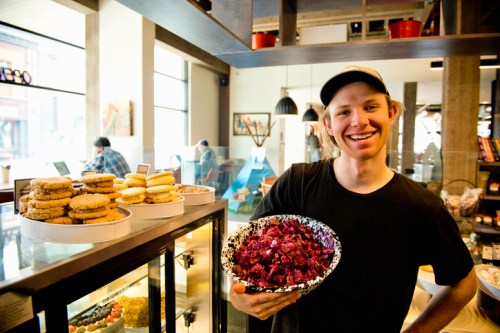 A smiling young man holds a bowl of colorful snacks in a cozy café, with cookies displayed in the background.