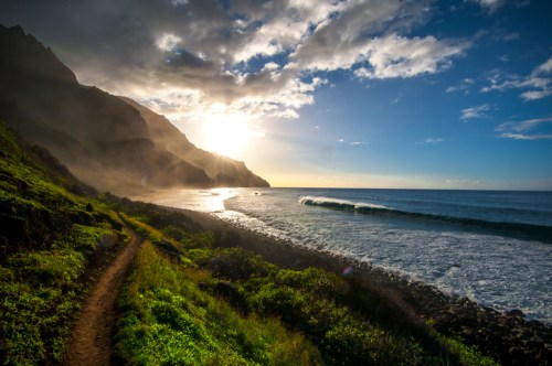 A scenic coastal view with waves, lush greenery, and mountains under a bright sky at sunrise.