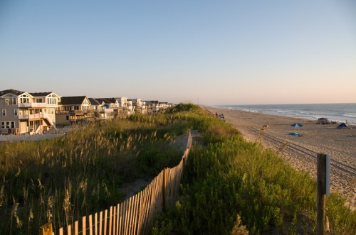 A serene beach scene at sunset, featuring dunes, beach houses, and gentle waves in the background.