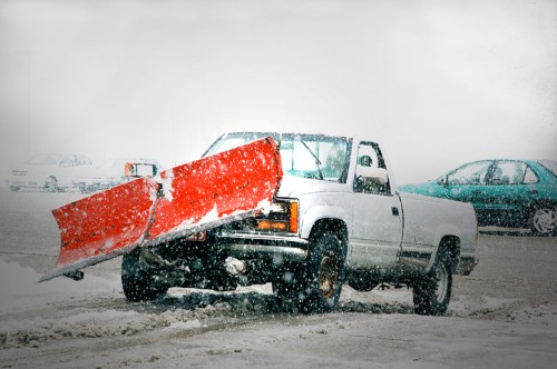 A white pickup truck with a red snow plow attachment parked in a snowy landscape during a snowstorm.
