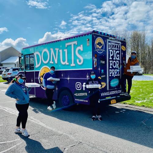 A colorful donut truck with four people wearing masks, holding boxes of donuts, against a sunny sky.
