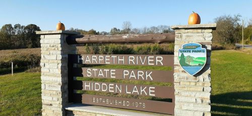 Sign for Harpeth River State Park, Hidden Lake, with pumpkins on top, surrounded by greenery and a clear sky.