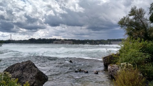 A turbulent river flows past rocky shores under a cloudy sky, with buildings visible in the background.