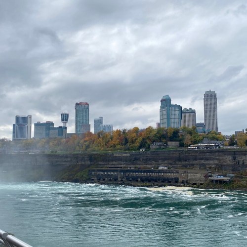 View of Niagara Falls with a skyline of tall buildings and autumn foliage along the riverbank under a cloudy sky.