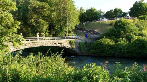 A stone bridge crosses a river, surrounded by lush greenery and people walking along a path in the background.