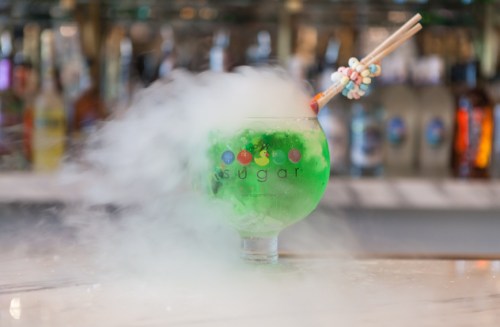 A vibrant green cocktail in a glass, topped with colorful candy and surrounded by mist.