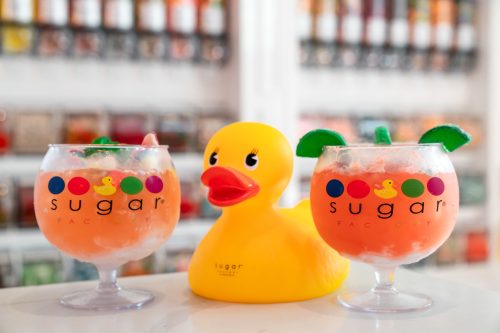 Two colorful cocktails with fruit garnishes sit beside a large rubber duck, all branded with "sugar" logos.