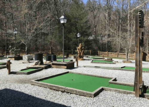 A mini-golf course with green turf, surrounded by trees and wooden fences, featuring a large statue in the background.