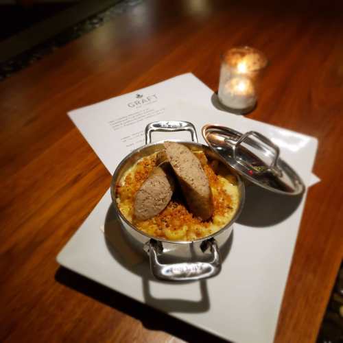 A dish of creamy macaroni and cheese topped with two sausage slices, served in a small pot on a wooden table.
