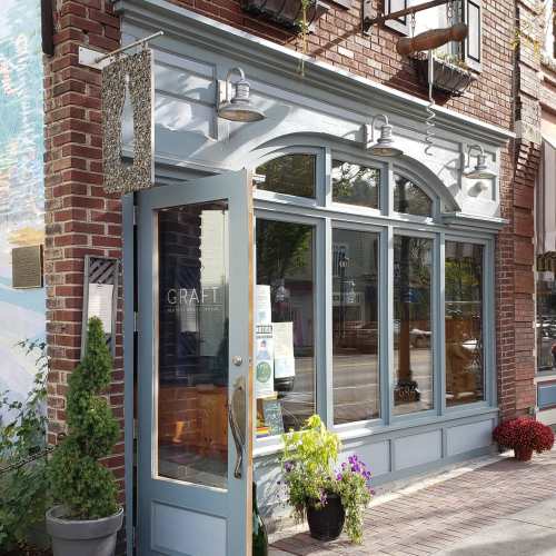 A storefront with large windows, featuring a sign for "GRAFT," surrounded by potted plants and brick walls.