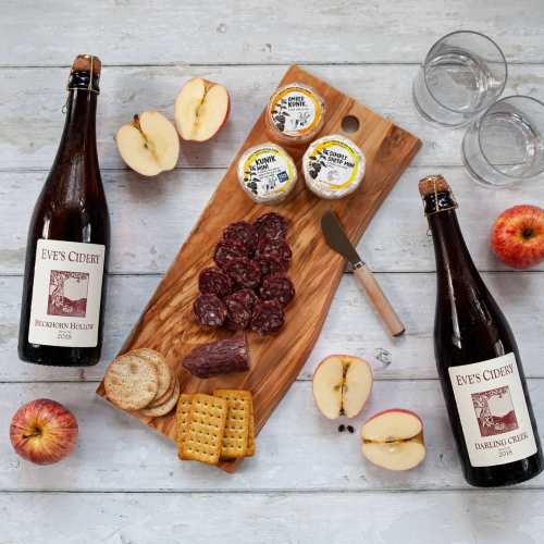 A wooden board with cheese, salami, crackers, apples, and two bottles of cider on a rustic table.