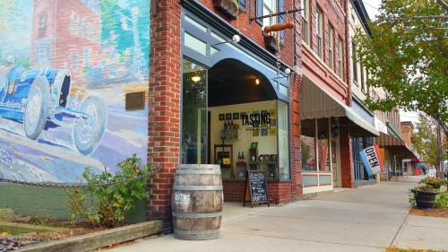 A brick building with a mural and an open tasting room, surrounded by shops along a tree-lined street.
