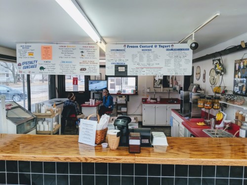 Interior of a dessert shop with a menu board, counter, and two customers seated at a table.