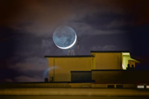 A crescent moon rises behind a building, partially obscured by clouds in a night sky.