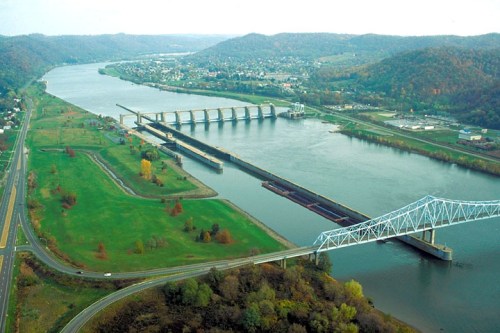 Aerial view of a river with a dam, barge, and a bridge, surrounded by green hills and trees.