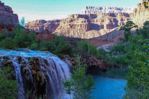 A serene waterfall cascades into a turquoise pool, surrounded by lush greenery and rugged canyon walls.