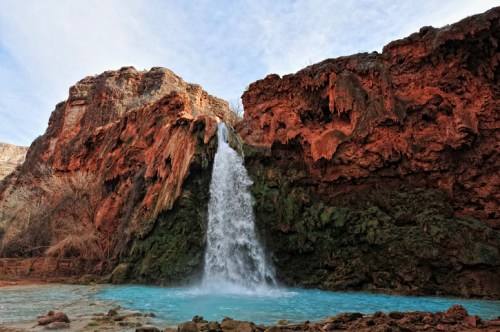 A stunning waterfall cascades over red rock cliffs into a turquoise pool below, surrounded by rugged terrain.