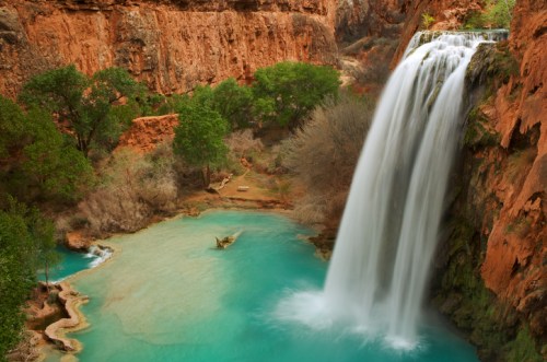 A stunning waterfall cascades into a turquoise pool, surrounded by red rock cliffs and lush greenery.