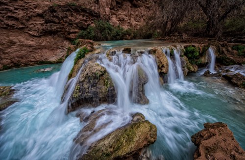 A serene waterfall cascades over rocks into a turquoise pool, surrounded by lush greenery and rugged canyon walls.