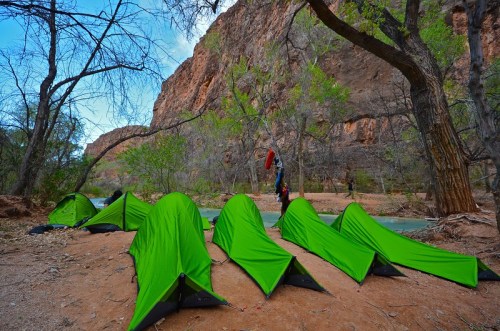 A row of bright green tents set up by a river, surrounded by rocky cliffs and sparse trees.