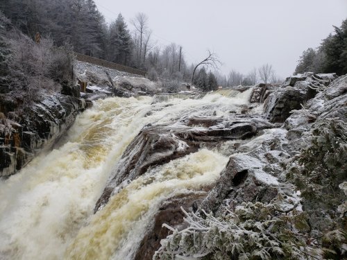 A snowy landscape featuring a rushing waterfall cascading over rocky terrain, surrounded by frosted trees.