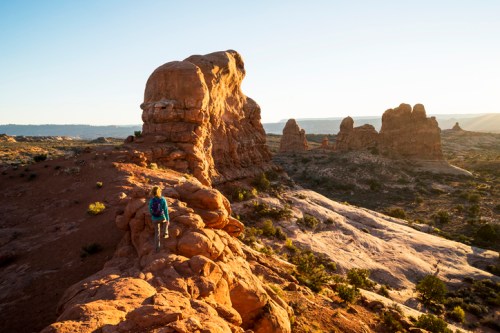 A hiker walks along a rocky trail in a desert landscape at sunset, with unique rock formations in the background.